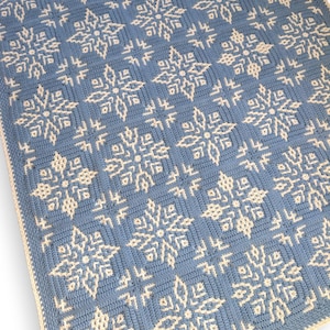May include: A handmade, crocheted blanket featuring a repeating snowflake pattern. The blanket is primarily light blue with off-white snowflakes. The edges are finished with a cream-colored border. The design is intricate and detailed, perfect for winter decor.