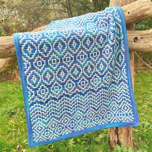 May include: A blue and white crocheted blanket with a geometric pattern. The blanket is draped over a wooden fence post.