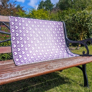 May include: A square crochet blanket featuring a repeating heart pattern in a gradient of purple and white. The design transitions from a deep purple border to a lighter, pale purple center, with white hearts integrated throughout the fabric.
