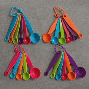 May include: Four sets of multi-coloured plastic measuring spoons, each set held together by a triangular ring. The spoons come in various sizes and vibrant colours including blue, orange, green, purple, pink, and red, arranged in a fan-like pattern.