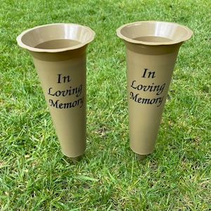 May include: Two olive-green memorial vases with flared tops and pointed bases are displayed on green grass. The vases have the words "In Loving Memory" printed in black script on the front.