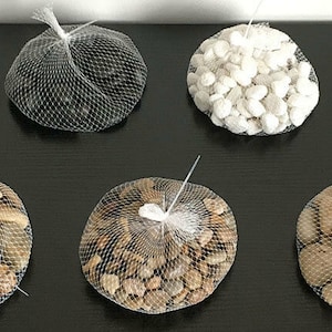 May include: Five circular mesh bags filled with decorative stones. One bag contains smooth black stones, one contains white stones, and three bags contain various shades of tan, brown, and grey river rocks. Each bag is tied at the top with a white tie.