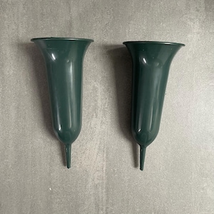 May include: Two dark green plastic vases with a flared top and a pointed base. The vases are empty and sit on a grey surface. The vases are likely for holding flowers.