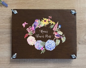 May include: A brown wooden flower press with a floral design and the text "Rina's Flower Press". The press has four metal screws on the corners.
