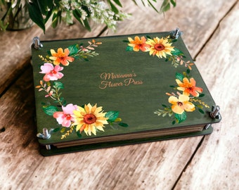 May include: A green wooden flower press with a floral design and the text "Marianna's Flower Press".