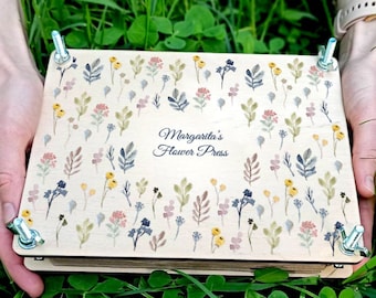 May include: A wooden flower press with a floral design and the text "Margarita's Flower Press." The press is held by hands in a bed of green grass. The floral pattern includes watercolor-style flowers in various colors.