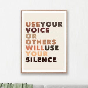 May include: A framed print with the text "USE YOUR VOICE OR OTHERS WILL USE YOUR SILENCE" in brown, orange, and black. The print is displayed above a grey sofa with decorative cushions and a green throw. A small white table is in front of the sofa.