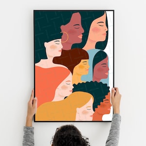 Peut inclure: Une affiche encadrée présentant une illustration colorée de visages de femmes diverses de profil. L'œuvre d'art présente une variété de tons de peau, de couleurs de cheveux et de styles, avec une esthétique minimaliste. L'affiche est tenue contre un mur blanc.