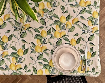 May include: A rectangular tablecloth featuring a repeating botanical print of yellow lemons and green leaves on a light beige background. A stack of two plain ceramic plates and a bowl, a silver fork, and a silver knife are placed on the fabric.