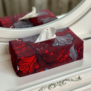 May include: A rectangular tissue box cover featuring a dark maroon fabric base with a printed pattern of large grey and red stylised leaves and floral elements. A single white tissue is pulled through the top opening of the box cover.