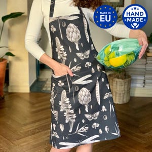 Könnte beinhalten: Schwarze Schürze mit botanischem Druck mit Artischocken, Libellen und Schmetterlingen. Die Schürze hat eine Tasche und verstellbare Träger. Handgefertigt und hergestellt in der EU.