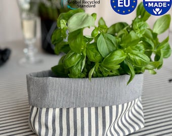 May include: A gray and white striped fabric basket filled with fresh green basil. The basket has a folded-over top and is sitting on a striped tablecloth. The image includes text that says "Global Recycled Standard", "Made in EU", and "Hand Made".