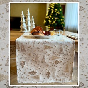 Könnte beinhalten: Cremefarbener Tischläufer mit einem sich wiederholenden Muster aus goldenen Weihnachtsbäumen und Sternen. Auf dem Läufer stehen ein Teller mit Gebäck und eine Tasse Kaffee. Dekorative silberne Weihnachtsbäume sind im Hintergrund.