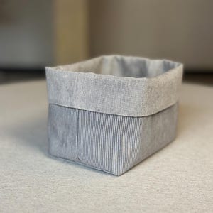 Op de afbeelding: Een rechthoekige stoffen opbergmand met een grijs en wit krijtstreepmotief aan de buitenkant. De bovenrand van de mand is omgeslagen om een contrasterende lichtgrijze, getextureerde stoffen voering aan de binnenrand te onthullen.