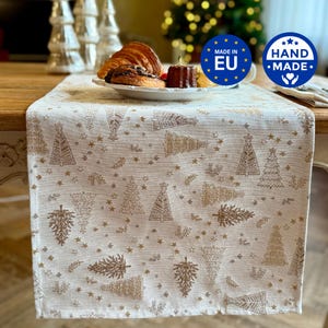 Op de afbeelding: Een rechthoekige tafelloper van witte stof met een herhalend patroon van goudkleurige kerstbomen en kleine sterretjes. Het ontwerp is over het gehele oppervlak gedrukt, wat zorgt voor een feestelijke uitstraling op de eettafel.