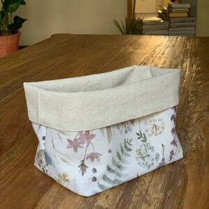 May include: A white fabric storage basket with a natural linen top. The basket features a floral pattern with watercolor-style flowers and leaves in shades of pink, green, and brown.