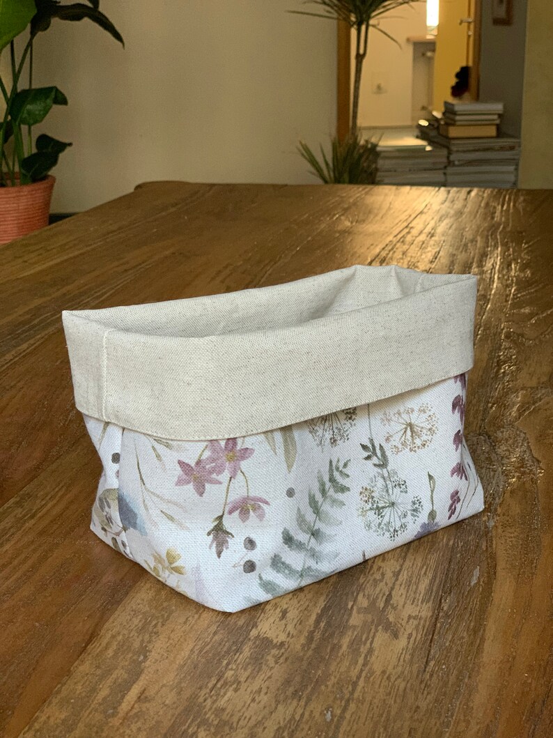 May include: A white fabric storage basket with a natural linen top. The basket features a floral pattern with watercolor-style flowers and leaves in shades of pink, green, and brown.