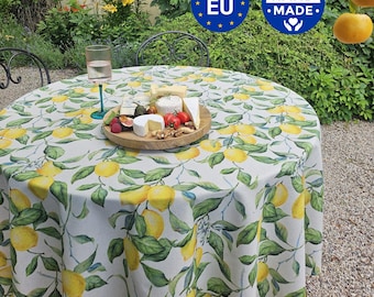 Può includere: Una tovaglia rotonda con un motivo ripetuto di limoni gialli e foglie verdi su sfondo bianco. Al centro si trova un tagliere in legno con vari formaggi, pomodorini e noci, accanto a un bicchiere alto.