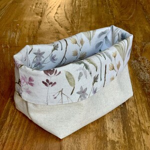 Op de afbeelding: Een stoffen opbergmand met een effen gebroken witte buitenkant en een omgeslagen bovenrand met een geprint patroon van delicate wilde bloemen, stengels en bladeren in gedempte tinten paars, groen en bruin op een lichte achtergrond.