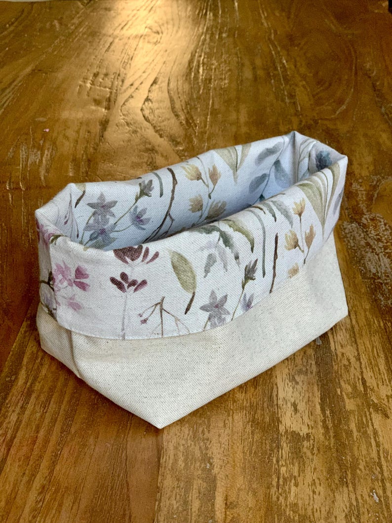 Op de afbeelding: Een stoffen opbergmand met een effen gebroken witte buitenkant en een omgeslagen bovenrand met een geprint patroon van delicate wilde bloemen, stengels en bladeren in gedempte tinten paars, groen en bruin op een lichte achtergrond.