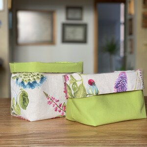 Op de afbeelding: Twee stoffen opbergmanden met een omgeslagen rand. E&eacute;n mand heeft een beige buitenkant met een kleurrijke botanische print van blauwe en roze bloemen en groene bladeren, gecombineerd met een effen limoengroene voering en bovenrand.