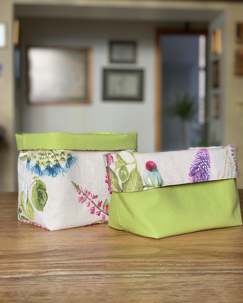 Op de afbeelding: Twee stoffen opbergmanden met een omgeslagen rand. E&eacute;n mand heeft een beige buitenkant met een kleurrijke botanische print van blauwe en roze bloemen en groene bladeren, gecombineerd met een effen limoengroene voering en bovenrand.