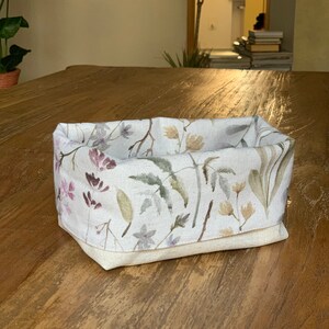 May include: A white linen storage basket with a floral pattern featuring pink, green, and brown watercolor leaves and flowers. The basket is sitting on a wooden surface.