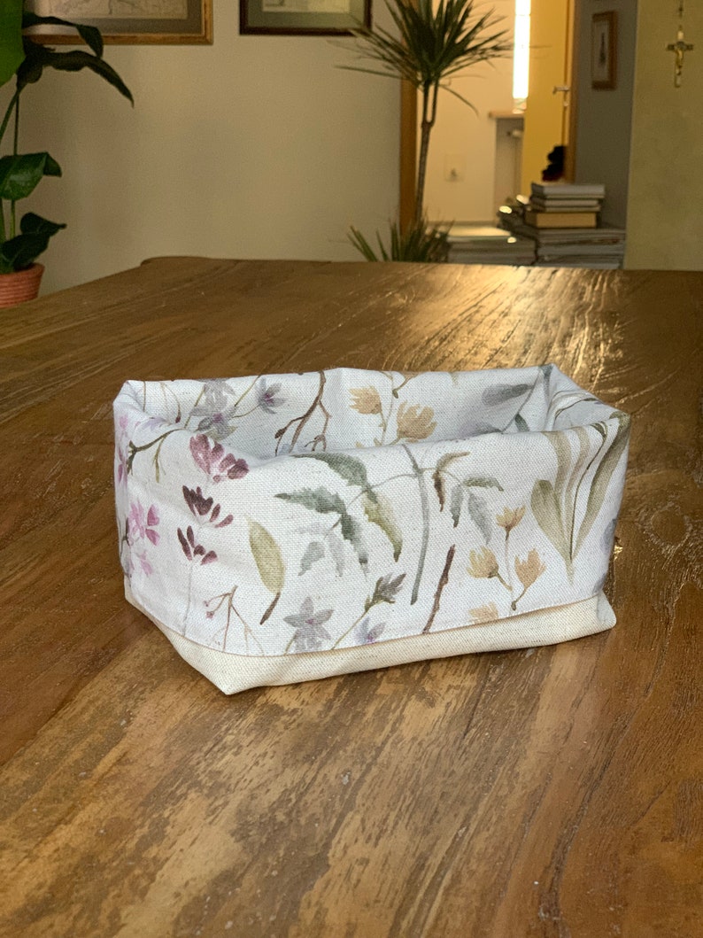 May include: A white linen storage basket with a floral pattern featuring pink, green, and brown watercolor leaves and flowers. The basket is sitting on a wooden surface.