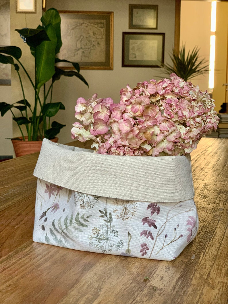May include: A white linen storage basket with a floral print featuring delicate green leaves and pink flowers. The basket is filled with dried pink hydrangeas.