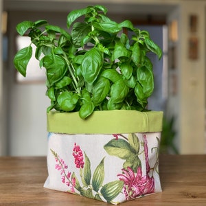 May include: A green fabric planter with a floral pattern featuring pink and green flowers. The planter is filled with a green basil plant.