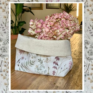 May include: A white linen storage basket with a floral print featuring delicate flowers and leaves in shades of pink, brown, and green. The basket is filled with a bouquet of dried pink hydrangeas.