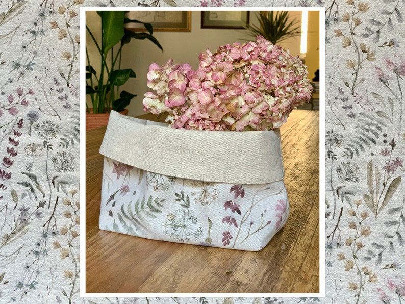 May include: A white linen storage basket with a floral print featuring delicate flowers and leaves in shades of pink, brown, and green. The basket is filled with a bouquet of dried pink hydrangeas.