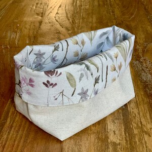 May include: A white fabric storage basket with a floral print featuring watercolor-style flowers in shades of pink, purple, and green. The basket is folded and sitting on a wooden surface.
