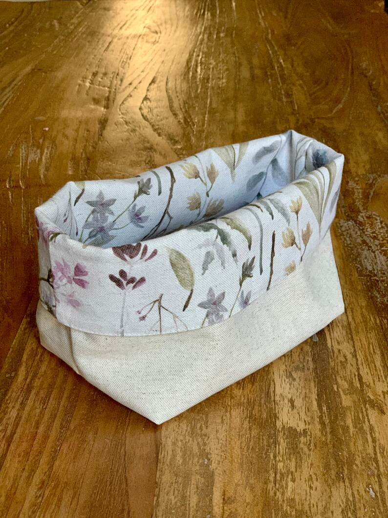 May include: A white fabric storage basket with a floral print featuring watercolor-style flowers in shades of pink, purple, and green. The basket is folded and sitting on a wooden surface.