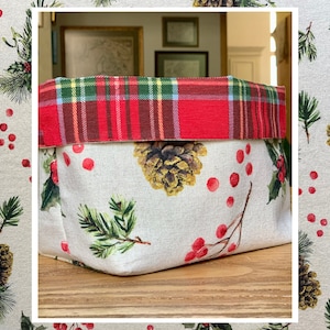 May include: A fabric storage basket with a festive Christmas theme. The basket features a white background with pine cones, holly, and red berries. The top edge is red plaid. The basket is sitting on a wooden surface.