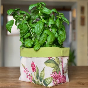 Op de afbeelding: Een vierkante stoffen plantenpotcover met een botanische print van rode bessen, groene bladeren en roze bloemen op een cr&egrave;mekleurige achtergrond. De bovenrand is afgewerkt met een effen limoengroene stoffen band, met een groene basilicumplant.