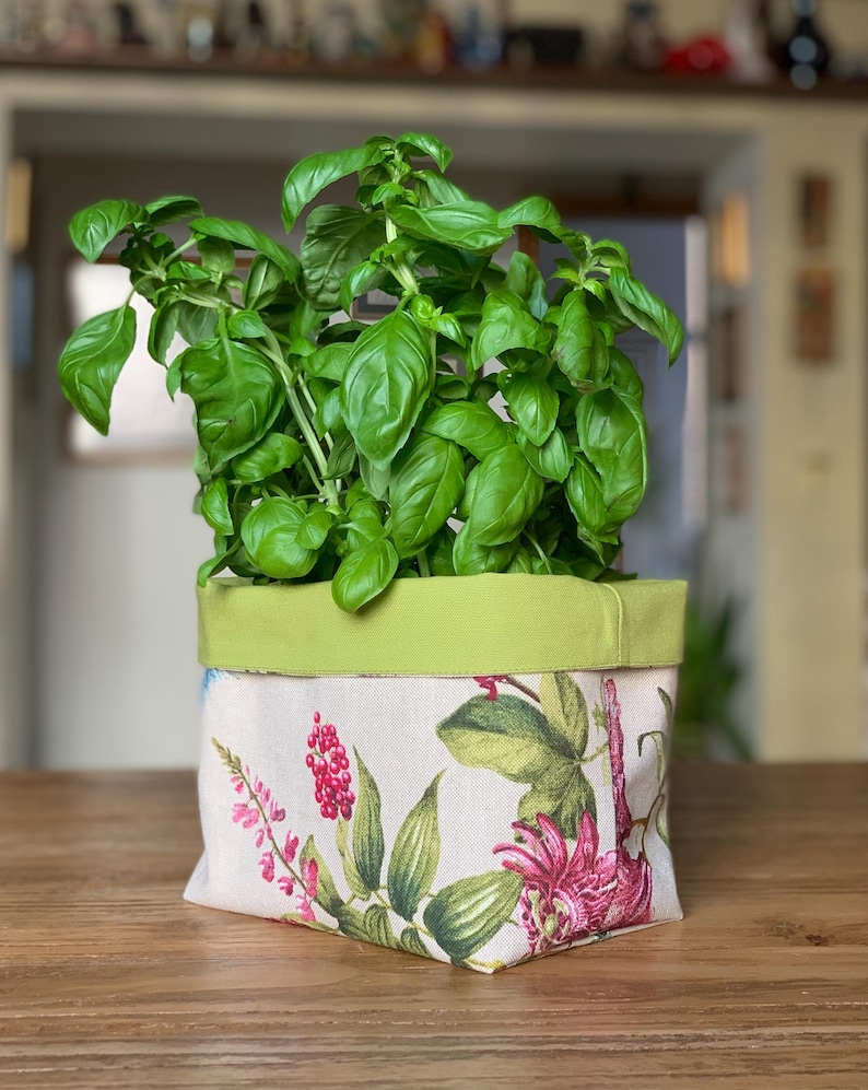 Op de afbeelding: Een vierkante stoffen plantenpotcover met een botanische print van rode bessen, groene bladeren en roze bloemen op een cr&egrave;mekleurige achtergrond. De bovenrand is afgewerkt met een effen limoengroene stoffen band, met een groene basilicumplant.
