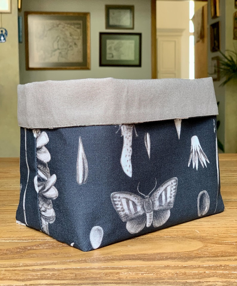 Op de afbeelding: Een rechthoekige stoffen opbergmand met een zwarte buitenkant en een wit printpatroon van vlinders, zaden en botanische elementen. De mand heeft een brede, omgeslagen bovenrand in een effen lichtgrijze kleur en een gestructureerde bodem.
