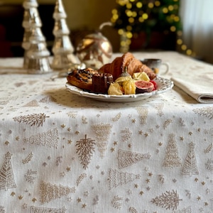 May include: A white tablecloth featuring an embroidered pattern of gold and silver Christmas trees and stars. The fabric has a textured weave and is draped over a table, serving as a base for a white plate filled with various pastries and fruit.