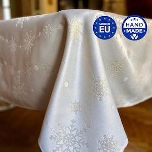 Könnte beinhalten: Eine weiße Tischdecke mit einem sich wiederholenden Muster aus silbernen Schneeflocken und dünnen Linien mit kleinen Kreisen. Oben rechts befinden sich zwei blaue runde Aufkleber, einer mit der Aufschrift MADE IN EU und einer mit HAND MADE und Herz.