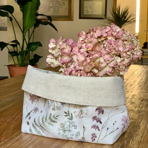 Op de afbeelding: Een stoffen opbergmand met een omgevouwen bovenrand in een natuurlijke beige kleur. Het onderste gedeelte is voorzien van een botanische print met aquarelachtige illustraties van diverse wilde bloemen, varens en grassen in gedempte roze, groene en bruine tinten.