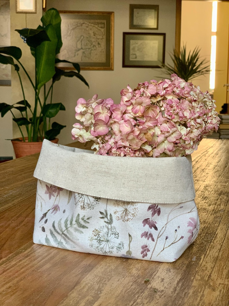 Op de afbeelding: Een stoffen opbergmand met een omgevouwen bovenrand in een natuurlijke beige kleur. Het onderste gedeelte is voorzien van een botanische print met aquarelachtige illustraties van diverse wilde bloemen, varens en grassen in gedempte roze, groene en bruine tinten.