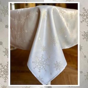 May include: White tablecloth with a snowflake pattern. The fabric drapes over a table, showcasing the intricate design. The snowflakes are in a light silver color, creating a festive and elegant look. Ideal for holiday or winter-themed events.