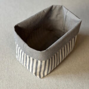 Op de afbeelding: Een rechthoekige stoffen opbergmand met een verticaal gestreept patroon in grijstinten en gebroken wit. De mand heeft een gestructureerd ontwerp met een omgevouwen bovenrand die een contrasterende voering met fijne grijze strepen onthult.