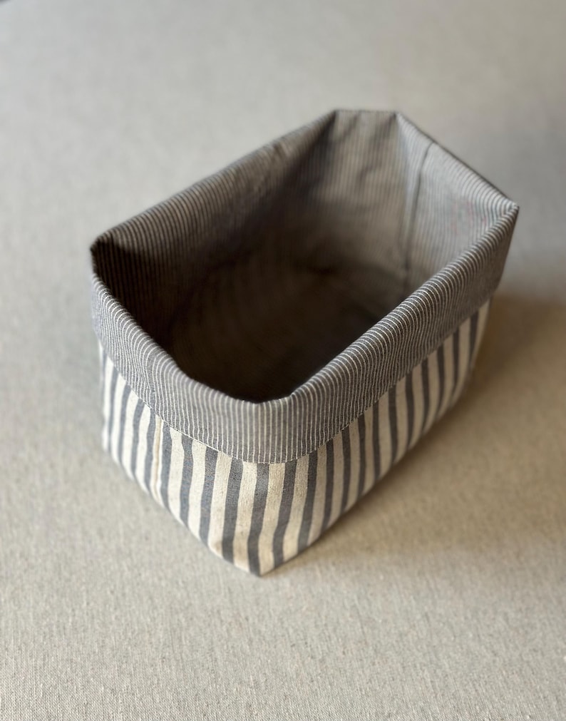 Op de afbeelding: Een rechthoekige stoffen opbergmand met een verticaal gestreept patroon in grijstinten en gebroken wit. De mand heeft een gestructureerd ontwerp met een omgevouwen bovenrand die een contrasterende voering met fijne grijze strepen onthult.