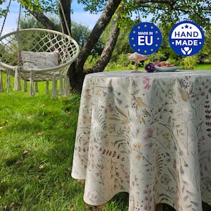 Könnte beinhalten: Eine runde Tischdecke mit einem botanischen Druck aus Wildblumen, Gräsern und Blättern in gedeckten Rosa-, Braun- und Blautönen auf cremefarbenem Grund. Der Stoff liegt auf einem runden Tisch, daneben ist ein hängender Makramee-Stuhl zu sehen.