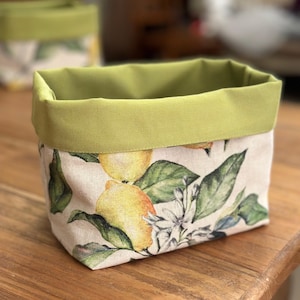 May include: A rectangular fabric storage basket featuring a pattern of yellow lemons, green leaves, and white blossoms on a light beige background. The basket has a solid olive green fabric trim along the top edge and a structured rectangular shape.