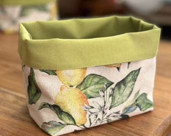 May include: A rectangular fabric storage basket featuring a pattern of yellow lemons, green leaves, and white blossoms on a light beige background. The basket has a solid olive green fabric trim along the top edge and a matching olive green interior lining.