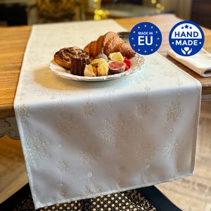 May include: A white table runner featuring a repeating pattern of light grey snowflakes and small circles. A white plate sits on the runner, holding a croissant, a dark pastry, a fig slice, and small yellow fruits. Two circular blue labels read MADE IN EU and HAND MADE.