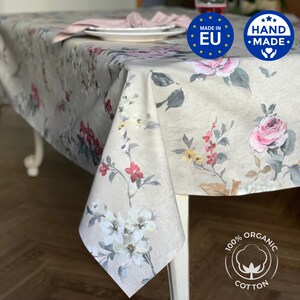 Op de afbeelding: Een beige bloemig tafelkleed met roze en rode rozen, witte en gele bloemen en groene bladeren. Het tafelkleed ligt op een witte tafel met een bord en een servet. De afbeelding bevat de tekst "HAND MADE" en "100% ORGANIC COTTON".